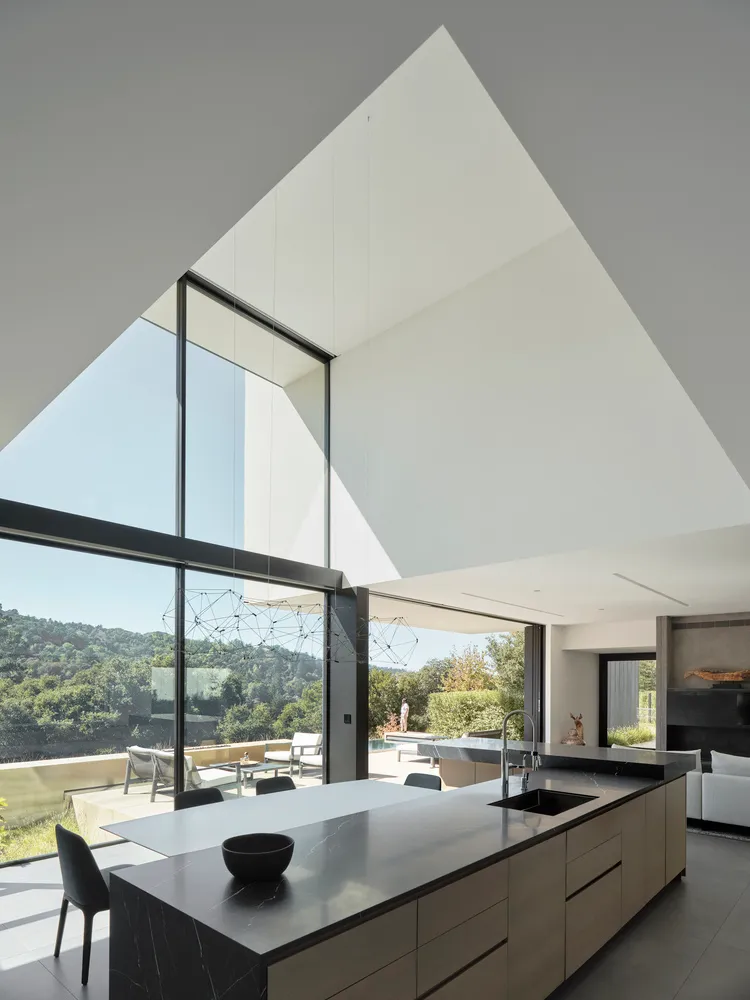 Modern kitchen with glass cabinets, open layout, and floor-to-ceiling windows overlooking greenery.