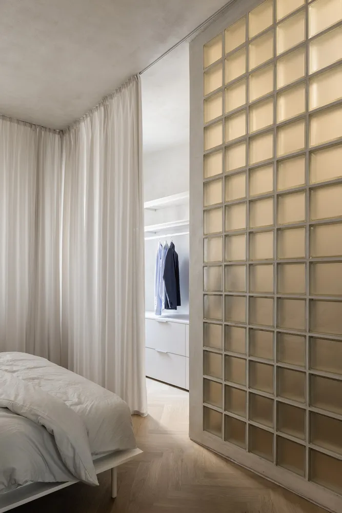 A minimalist bedroom featuring a glass-enclosed wardrobe, white furniture, and wooden flooring.