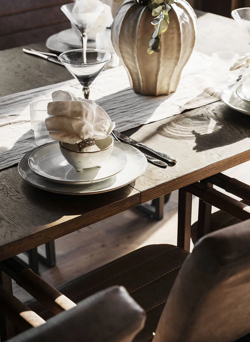A modern dining room featuring wood furniture, white plates, and a gold ring on a napkin, with a vase and place settings.
