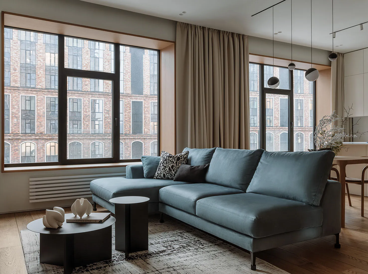 A modern living room featuring a blue sofa with patterned and dark pillows, black coffee tables, and a large window with beig