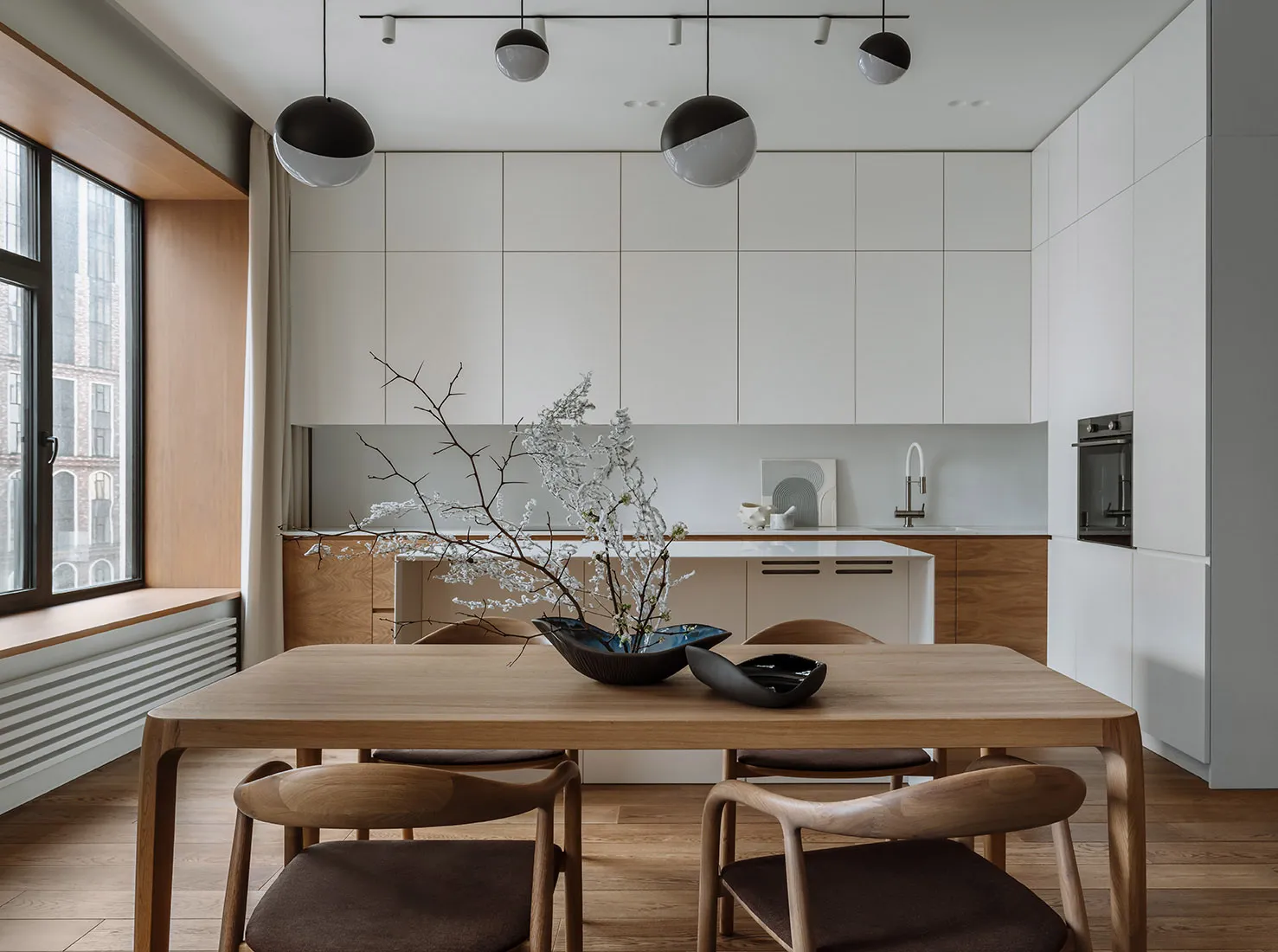 Modern minimalist kitchen with white cabinetry, wooden table and chairs, and a white backsplash with a decorative plant arran