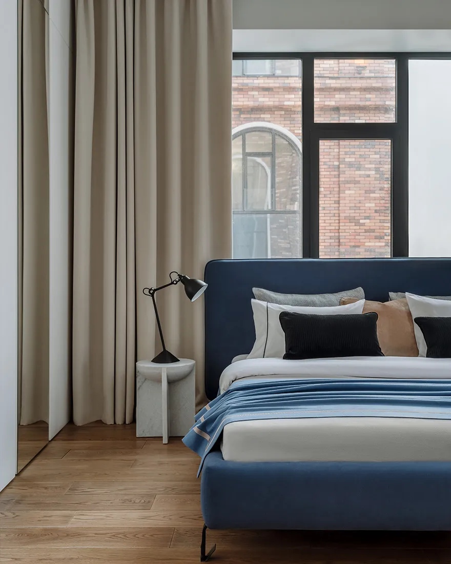 A minimalist bedroom with a blue upholstered bed, beige curtains, and a wooden floor.