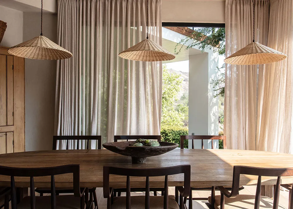 A minimalist dining room with wooden furniture, three wicker pendant lights, and long curtains.