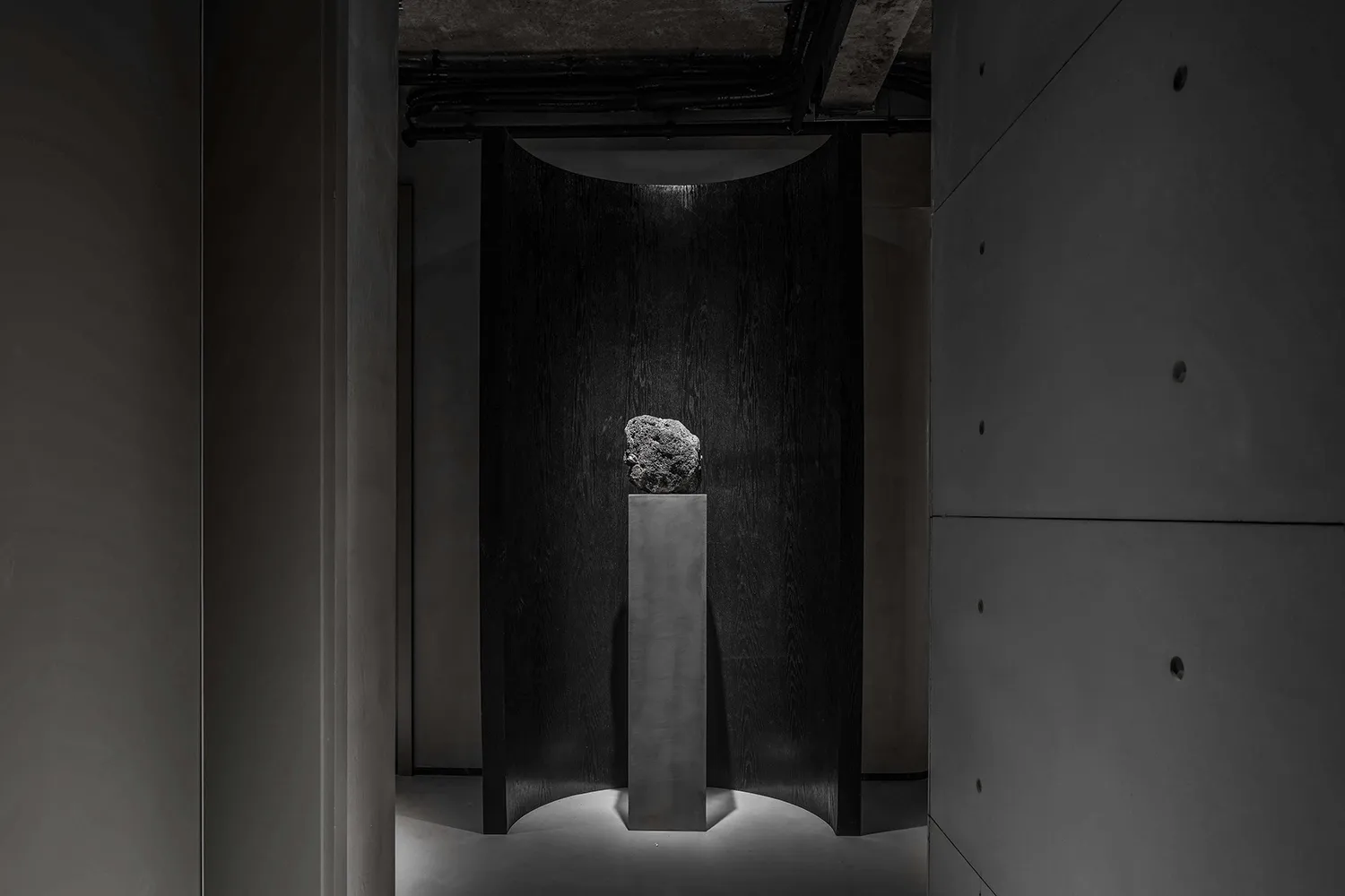 A minimalist exhibition space featuring a metal monolith against a dark background.
