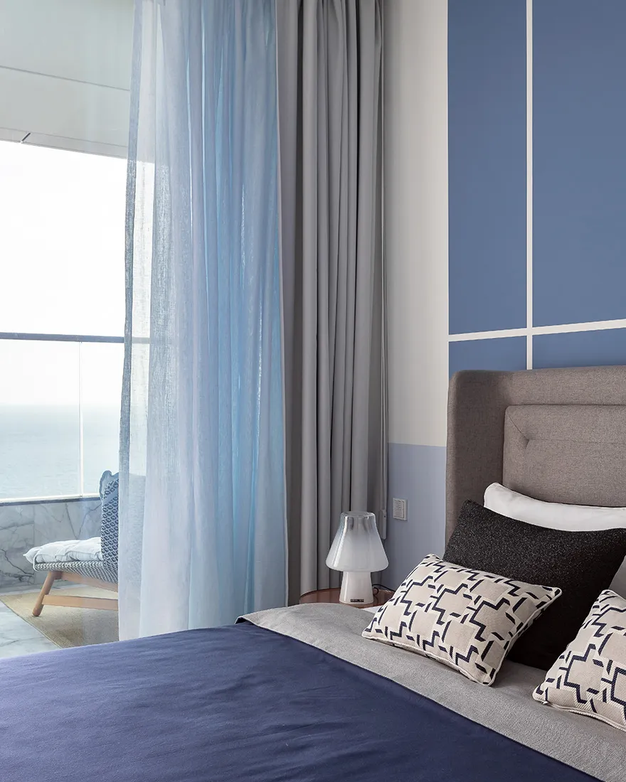 A minimalist bedroom featuring gray and blue textiles, with a view of the ocean from the balcony.