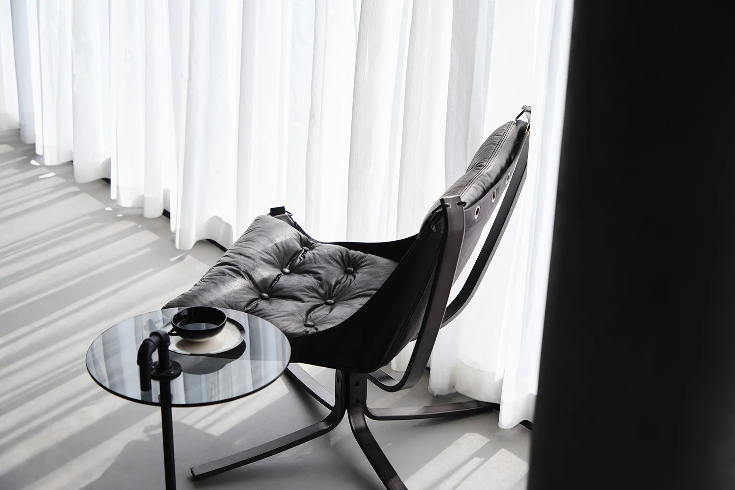 A sleek, modern leather chair with white cushions sits next to a round glass table with a mug and plate, against a backdrop o