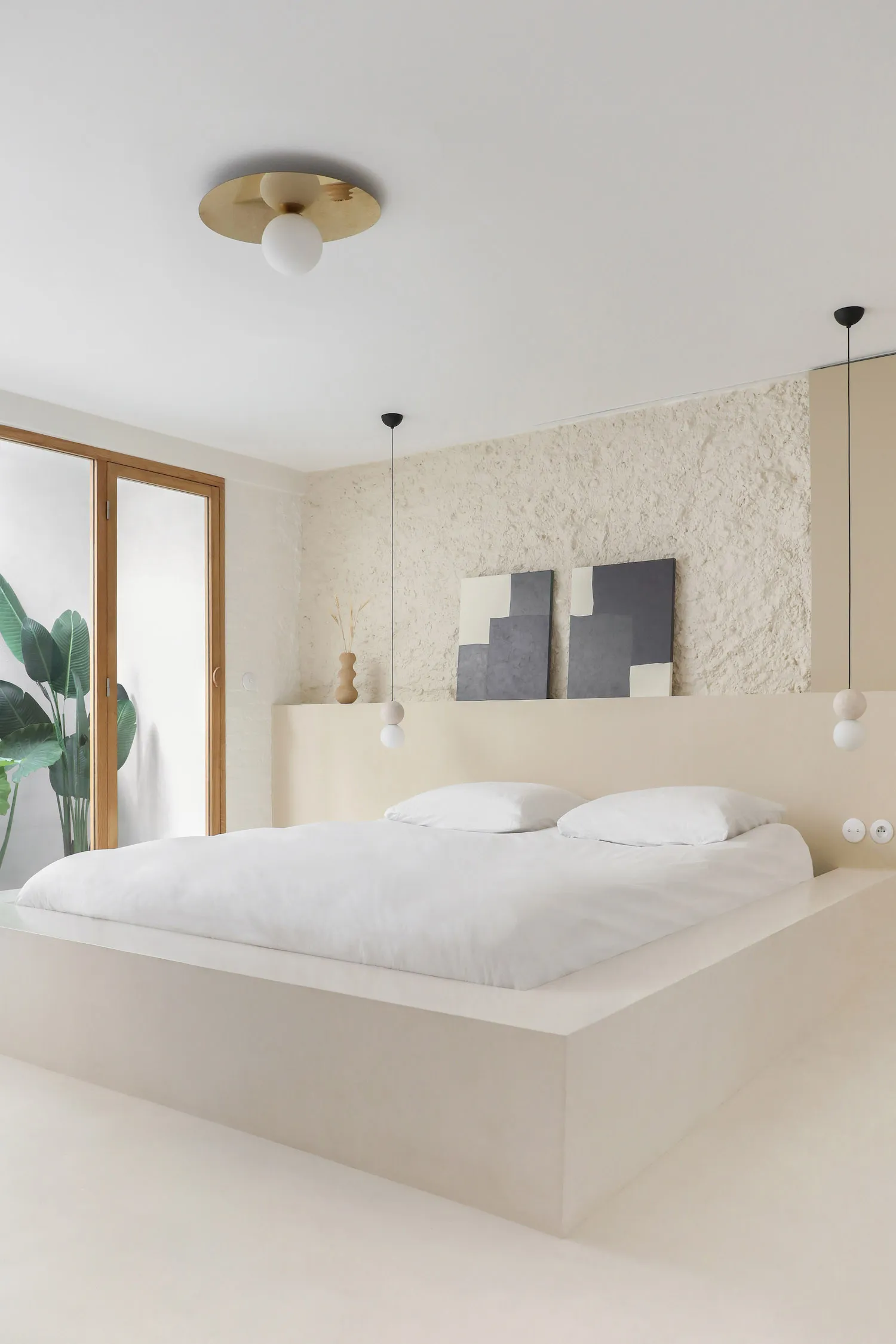 A minimalist bedroom with mixed materials, featuring a large bed with white bedding, a textured wall, and hanging light fixtu