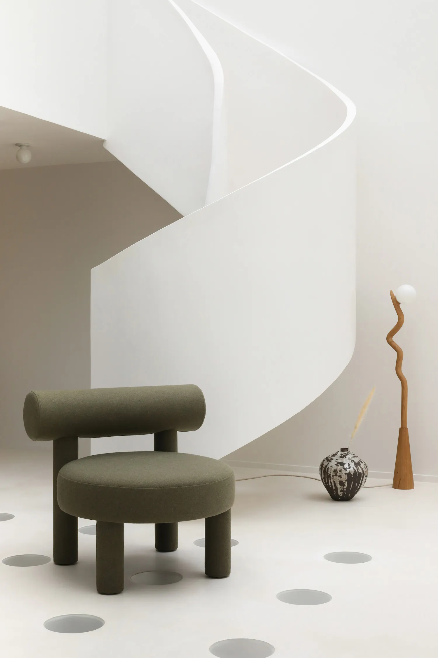 A modern green upholstered armchair with round legs, situated in a minimalist white interior space with a curved white wall a