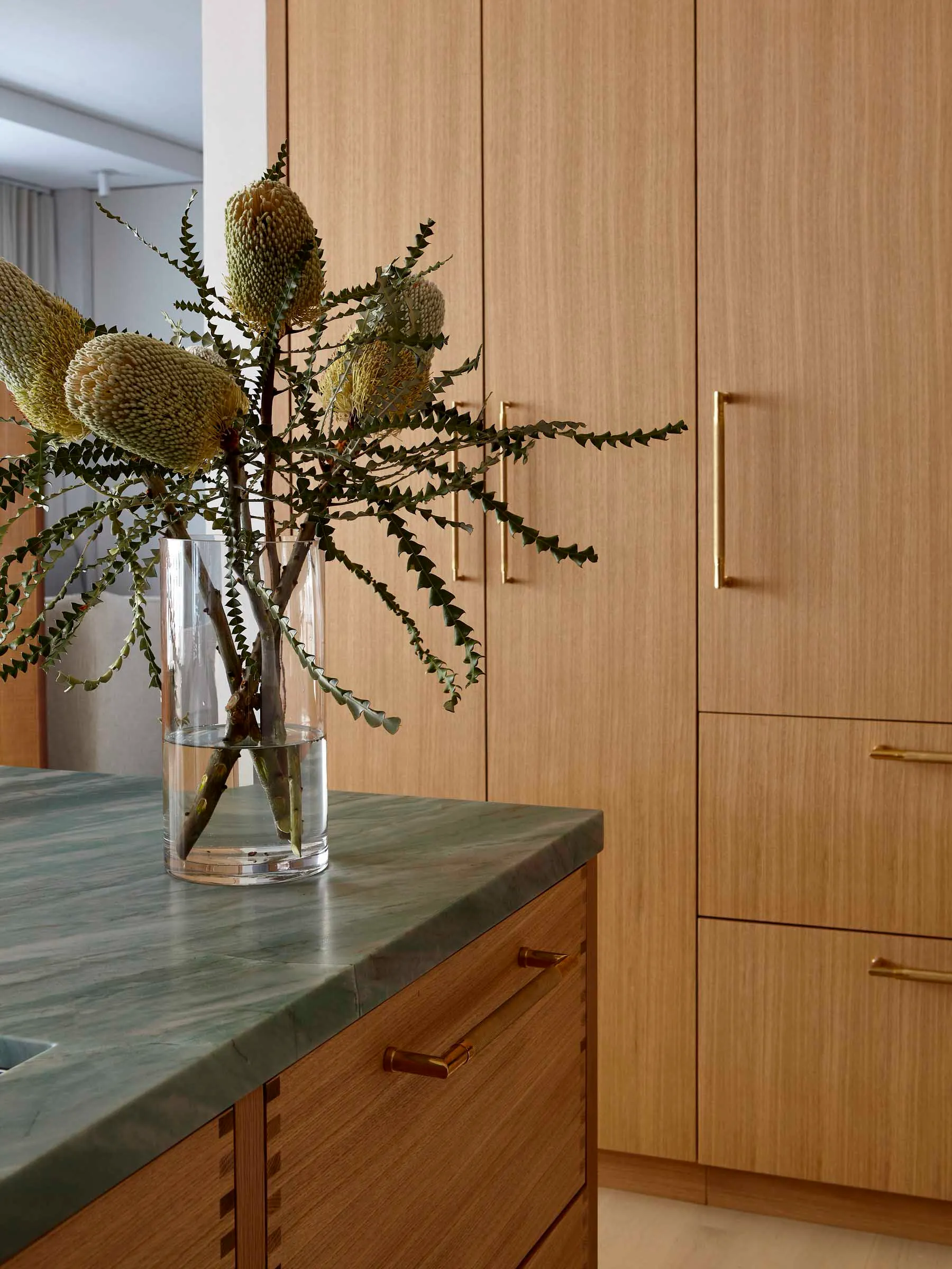Modern minimalist kitchen with wooden cabinets and marble countertop featuring Banksia flowers in a vase.