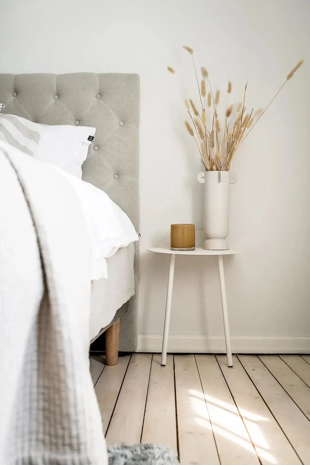 A minimalist bedroom with a fabric bed against a white wall, a white side table with a vase of dried plants and a candle hold