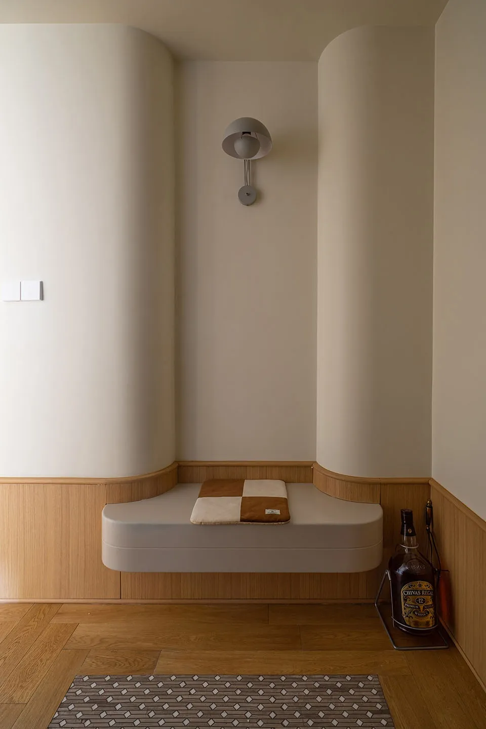 A minimalist seating area with a wooden bench and cushion, featuring a wall-mounted lamp and a bottle of Chivas Regal whisky.