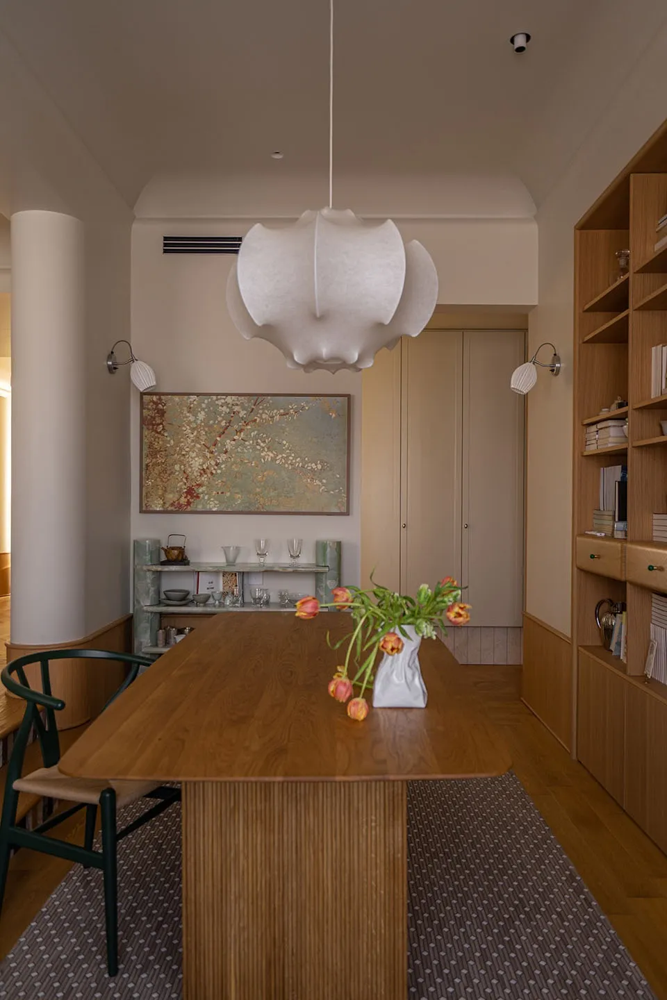 Modern minimalist dining room with wooden table, white pendant light, abstract painting, and bookshelf.