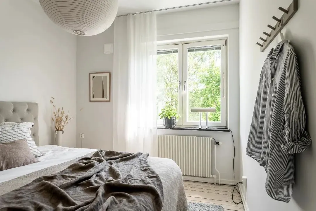 A minimalist bedroom featuring a gray blanket on a bed, a window with greenery outside, and a coat rack with a striped shirt.