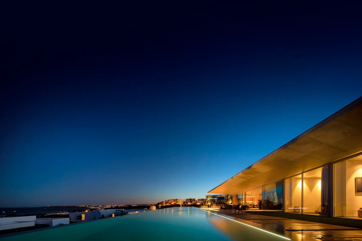 A modern residential home at night, featuring a glass and concrete exterior with an infinity pool.