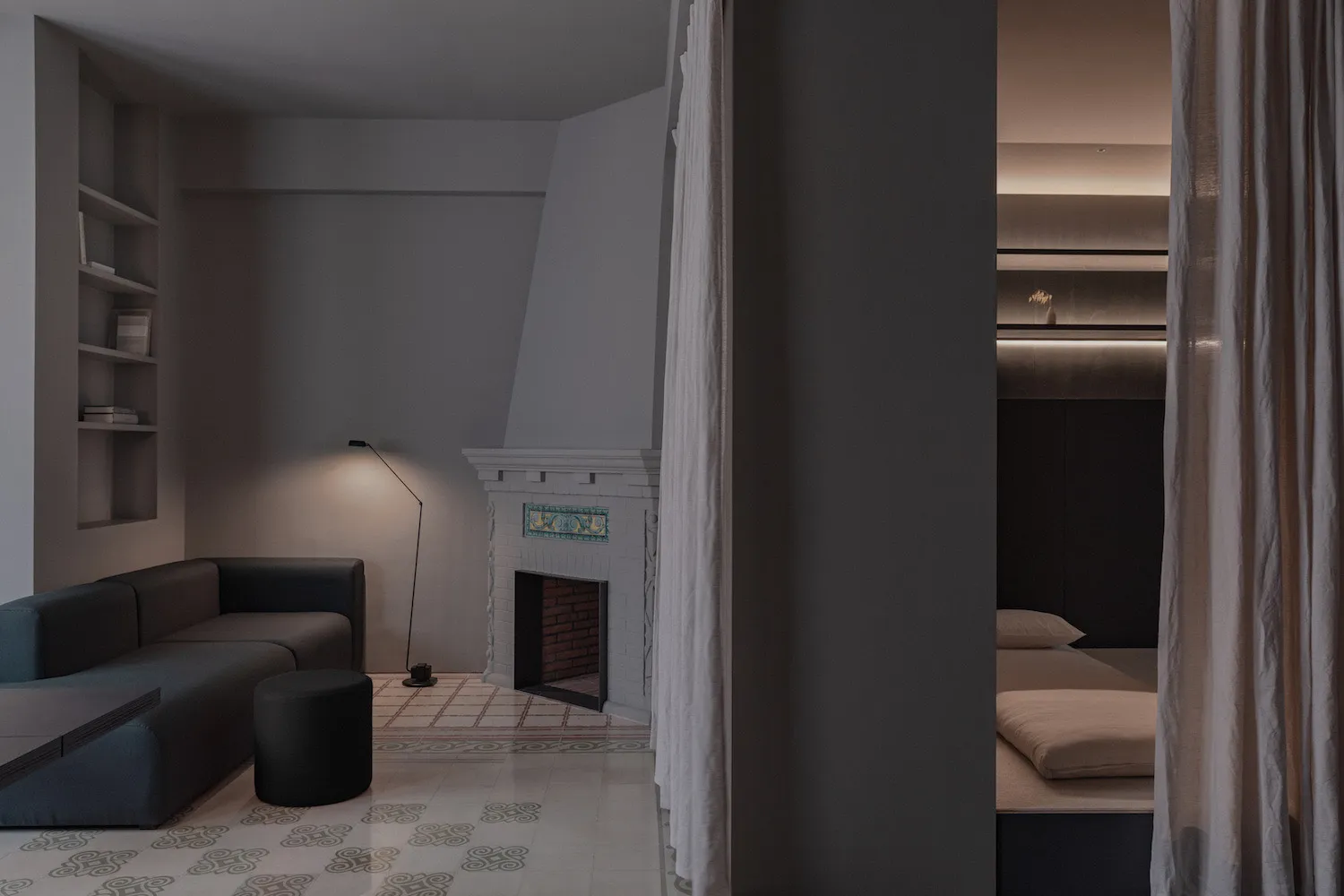 A minimalist bedroom featuring a plaster floor, a bed with beige bedding, and curtains leading to a separate sleeping area wi