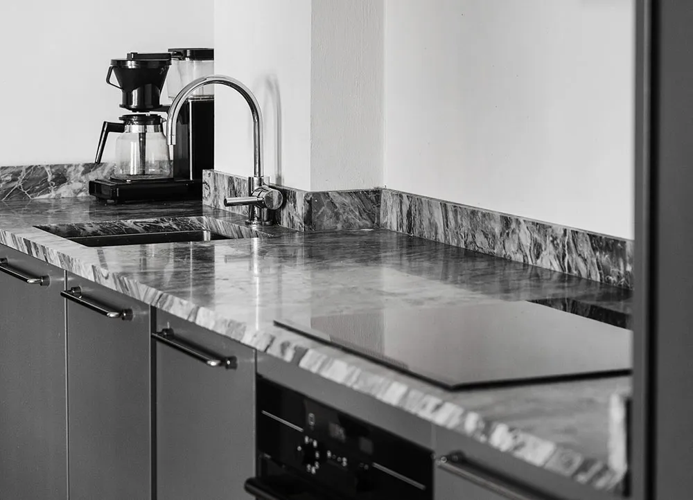 A modern minimalist kitchen featuring a marble countertop with a stainless steel sink and faucet, alongside a black coffee ma