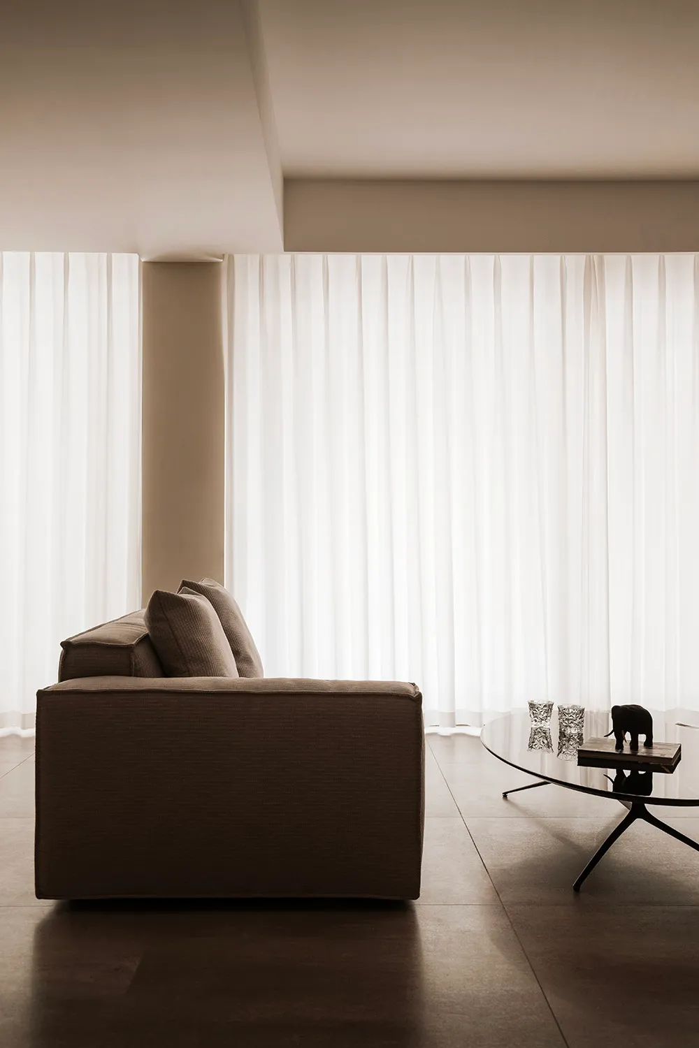 A minimalist living room with a brown upholstered sofa and white sheer curtains on the window, creating a serene and modern a