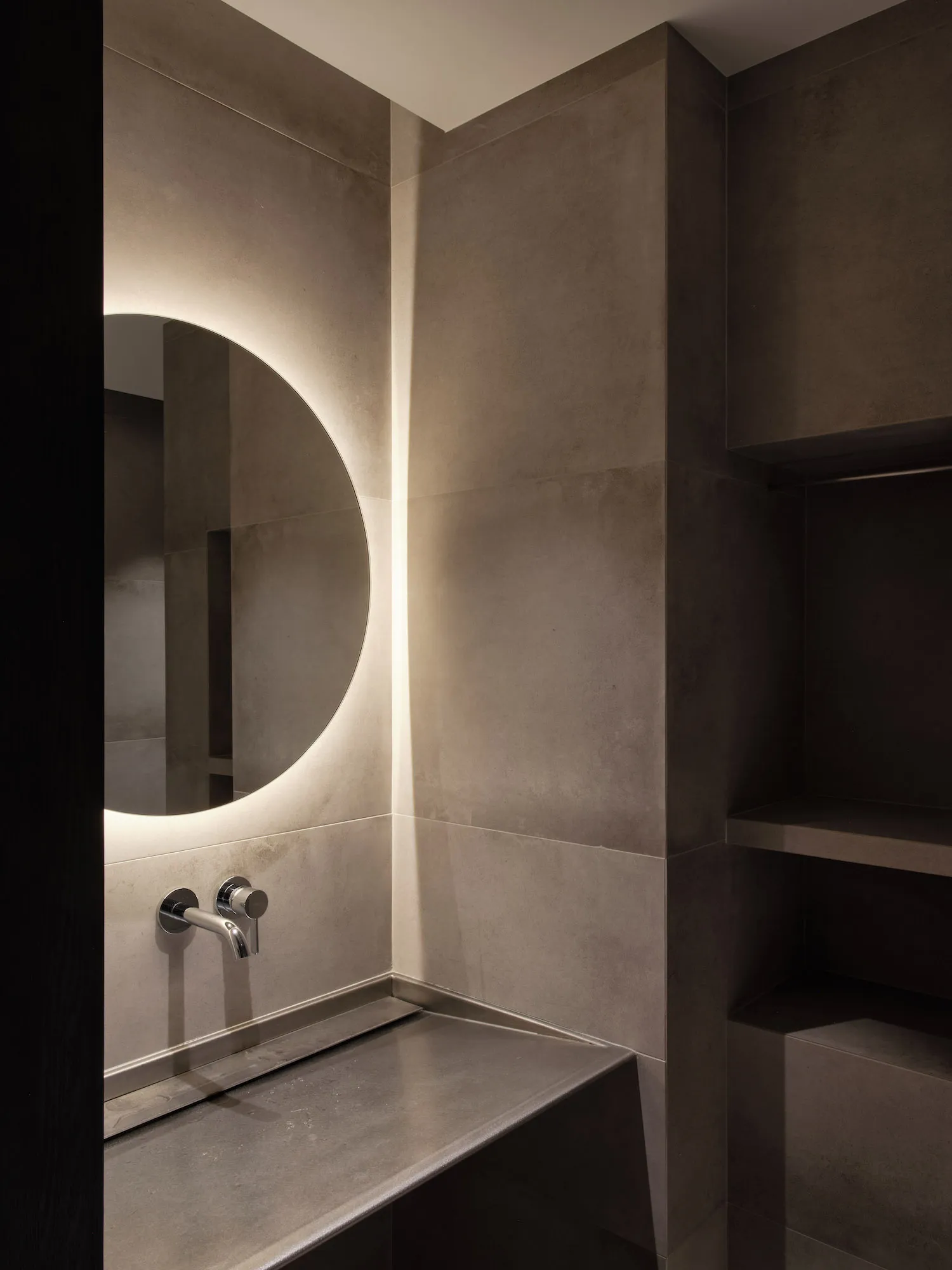 A modern bathroom interior with concrete walls and a round mirror with backlighting.