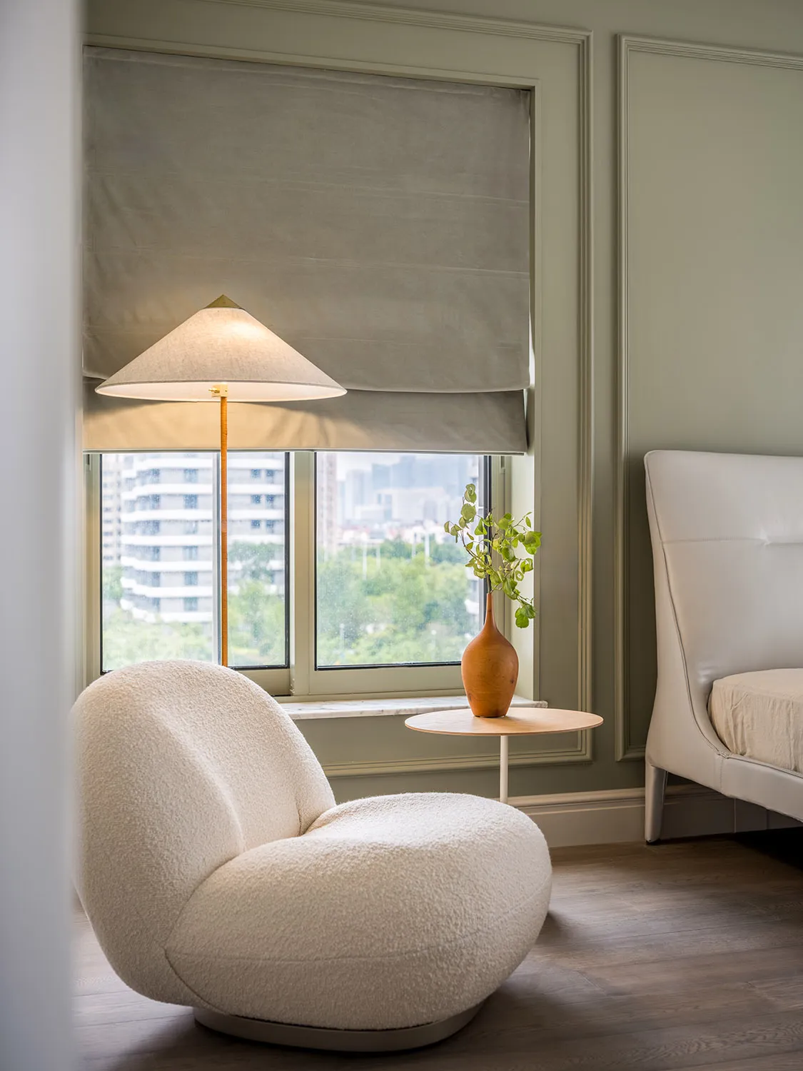 A modern bedroom featuring white upholstered furniture, a floor lamp, and a small round table with a vase containing greenery
