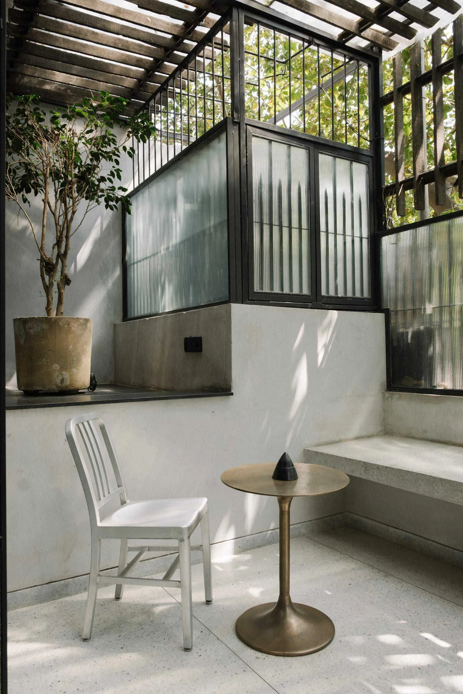 A modern industrial design space featuring a concrete floor, metal furniture, and natural light filtering through a pergola.