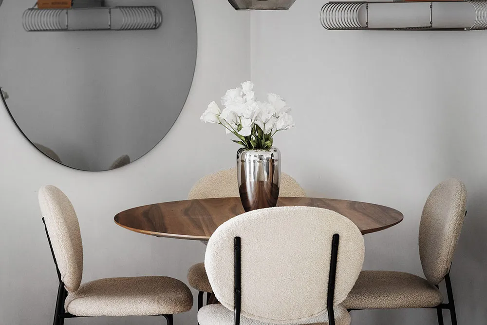 A modern dining room with wooden table, upholstered chairs, metallic accents, and a vase of white flowers.