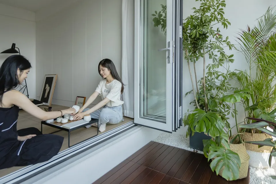 Two women sitting on a wooden floor, engaged in a tea ceremony surrounded by green indoor plants.