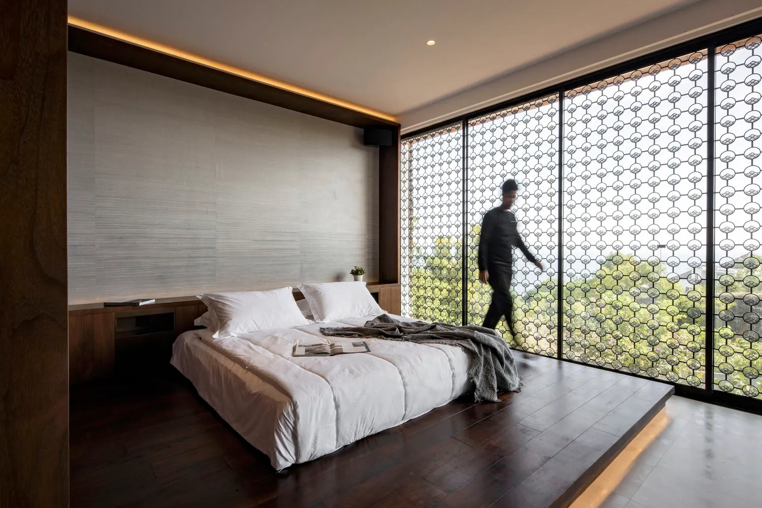 A modern minimalist bedroom with a concrete backdrop, featuring a large bed with white bedding and a wooden floor.