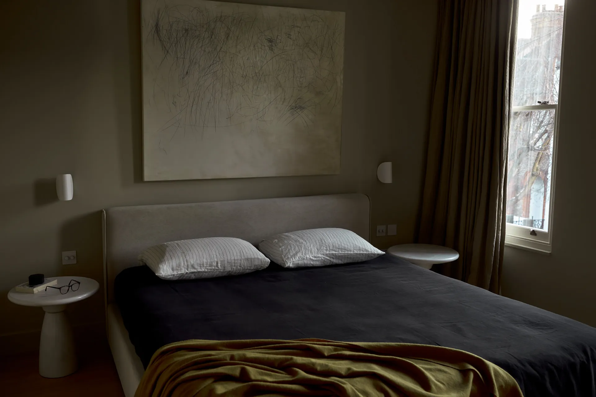 A neatly made bed with two white striped pillows and a dark blanket, featuring a large abstract artwork above it and a window