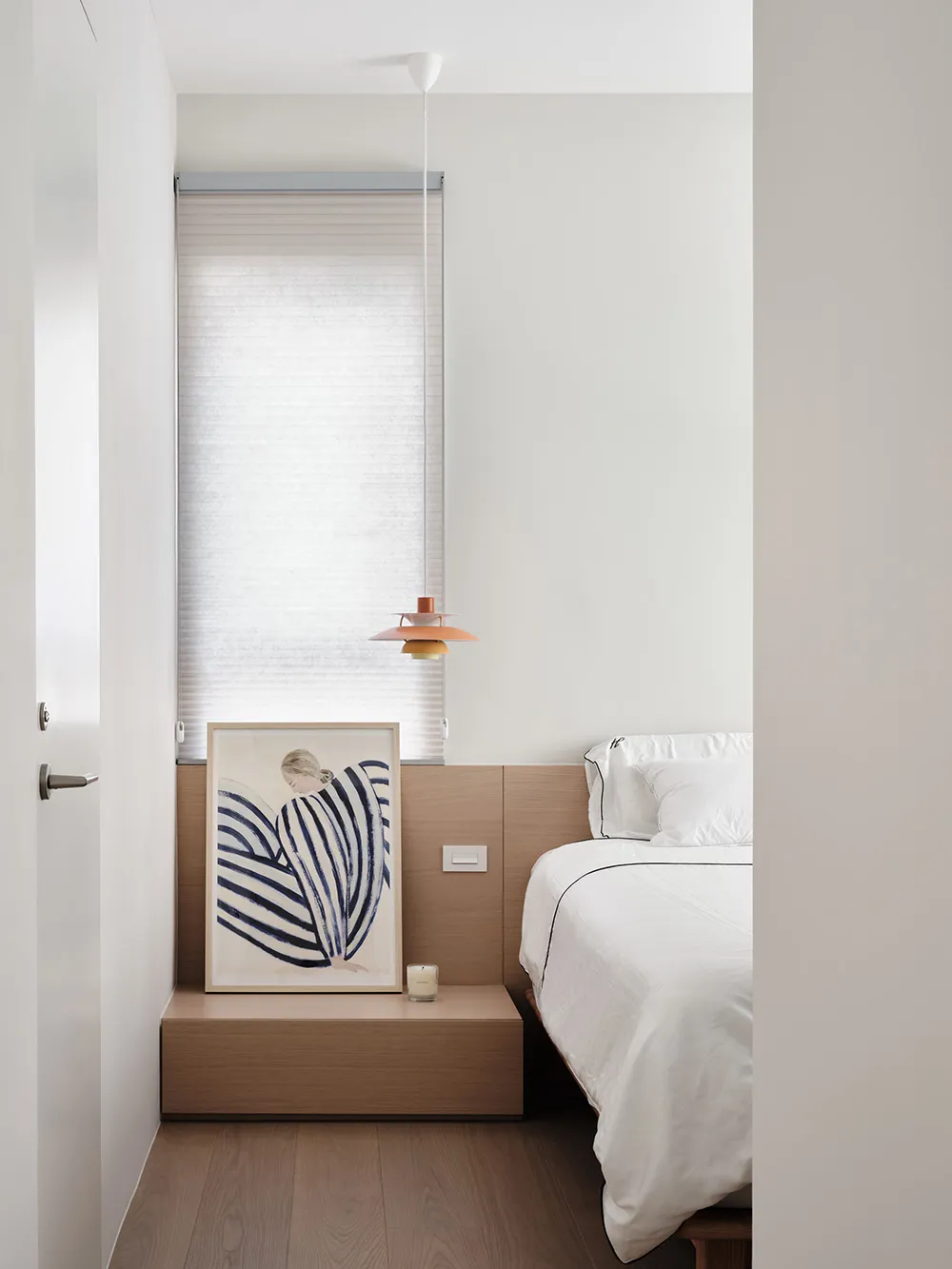 A minimalist bedroom with a wooden headboard, a white bed, and a framed abstract painting.