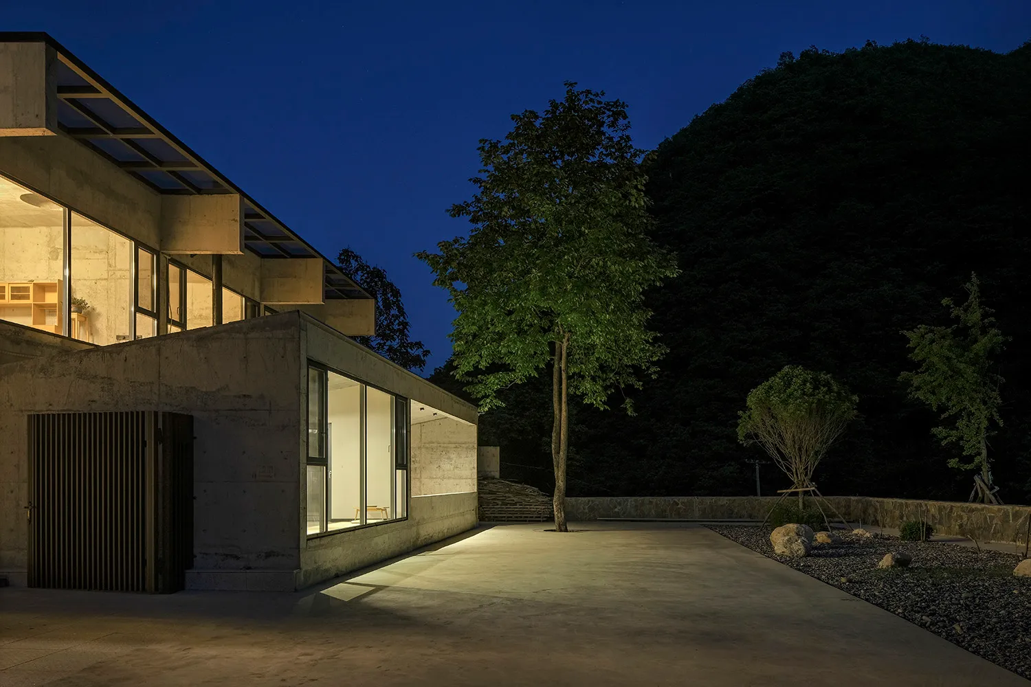 Residential building with concrete walls and glass windows illuminated at night, surrounded by a garden with trees and rocks.