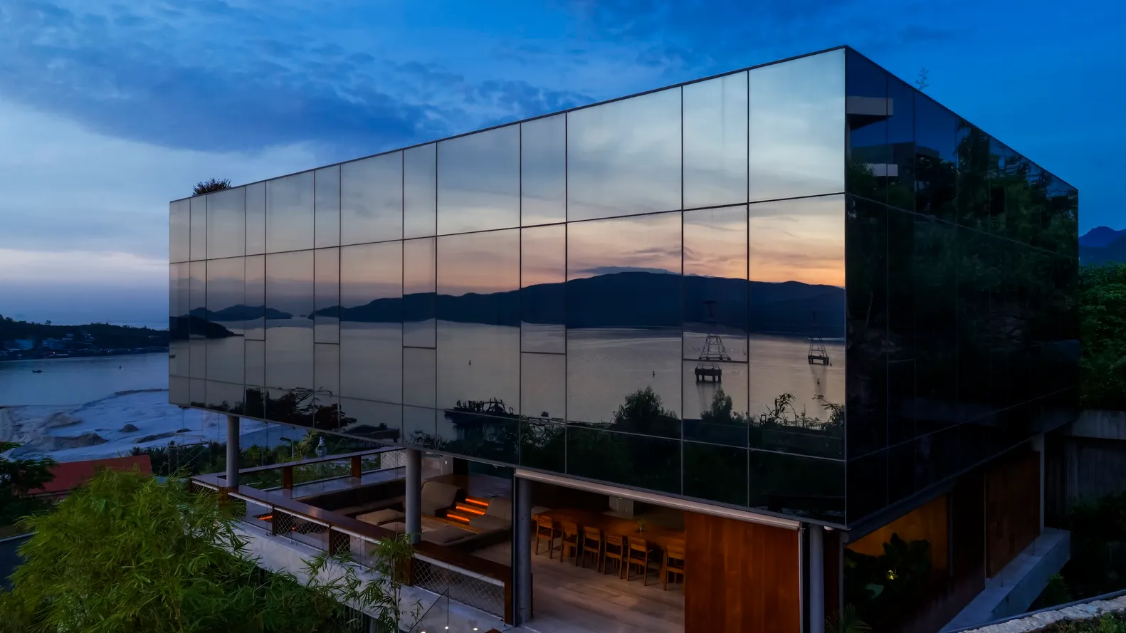 Modern residential house with glass windows reflecting a sunset and surrounding landscape.