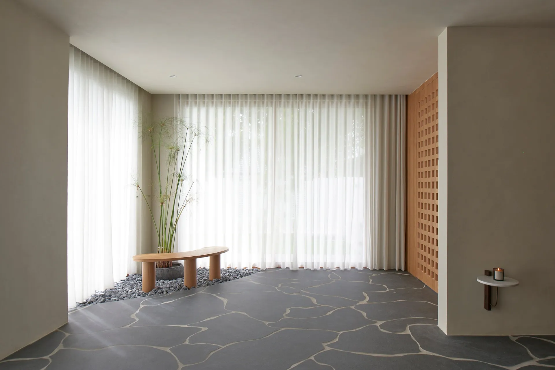A minimalistic living room featuring a grey, stone-patterned floor, a white sheer curtain, and a wooden bench seat with a pla