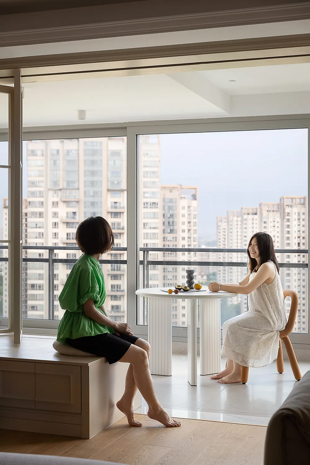 兩位女性坐在現代客廳的白色瓷磚地板上，眺望窗外城市景觀。