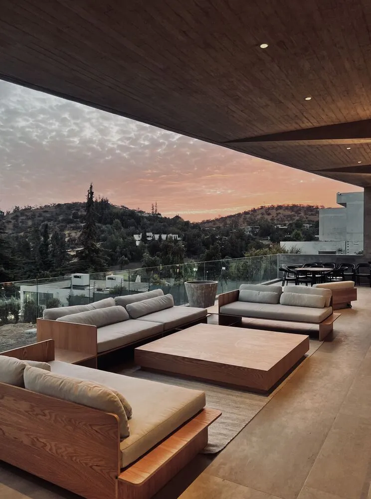A modern minimalist outdoor living space featuring wooden furniture and a view of a sunset over a hillside.