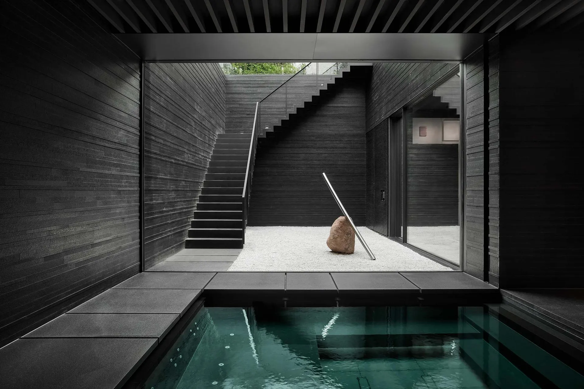 Modern minimalist interior space featuring concrete walls and floor, with a central rock sculpture and a glass staircase.