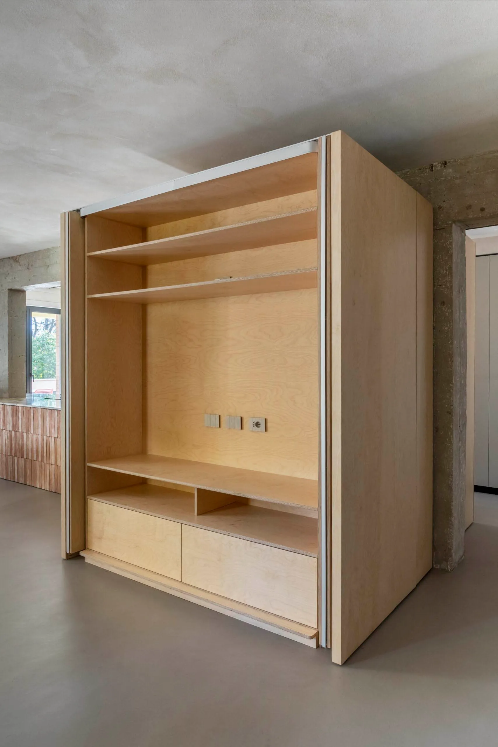 A minimalist birch plywood closet system with shelves and drawers in a clean, industrial room with concrete walls and a concr