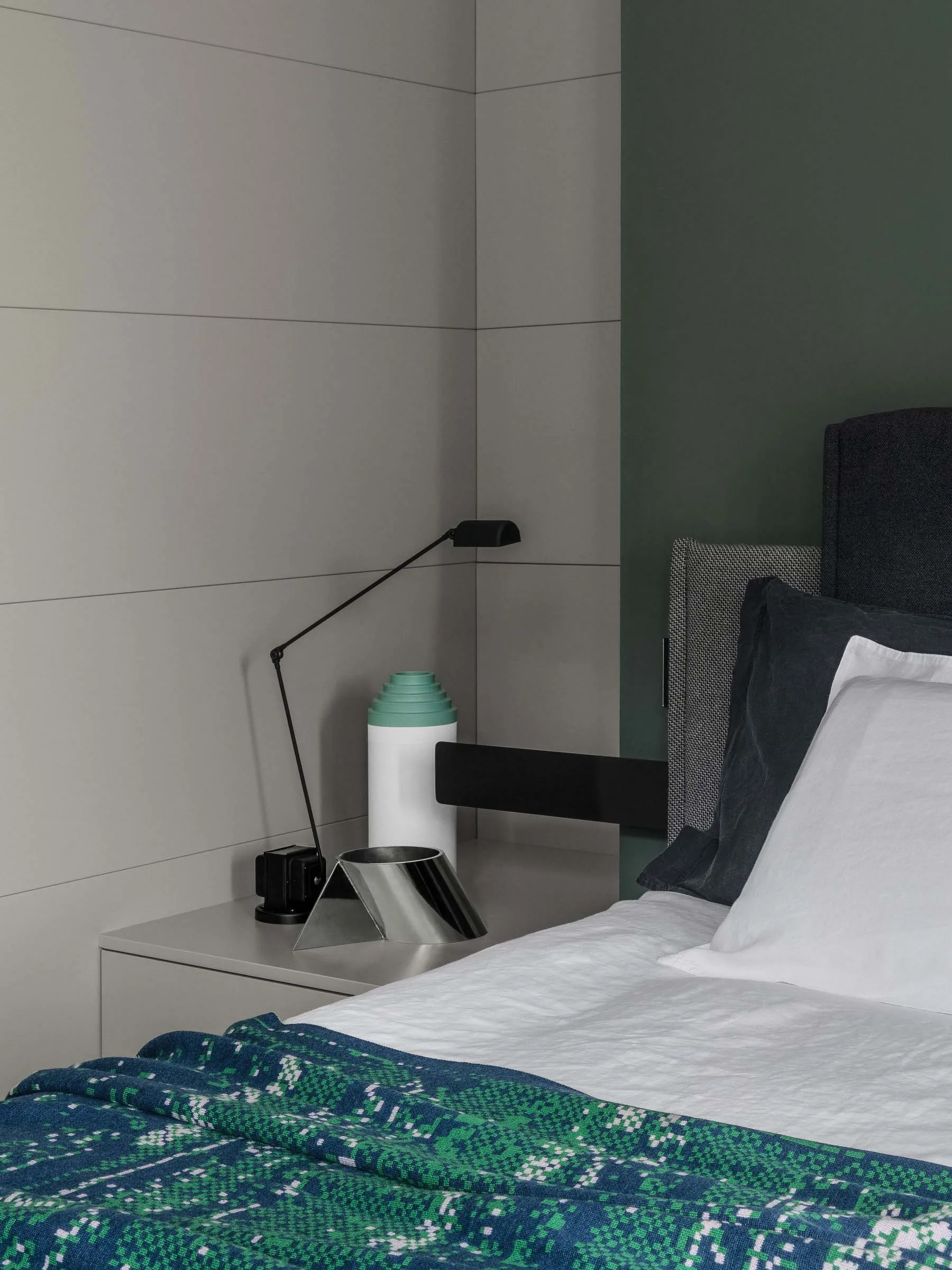 A minimalist bedroom with concrete, metal, and fabric materials, featuring a bed with a green patterned blanket, a modern lam