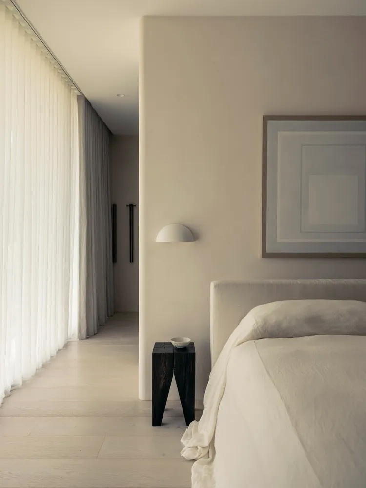 A minimalist bedroom featuring a white fabric upholstered bed, a wooden nightstand, and light gray curtains on a large window