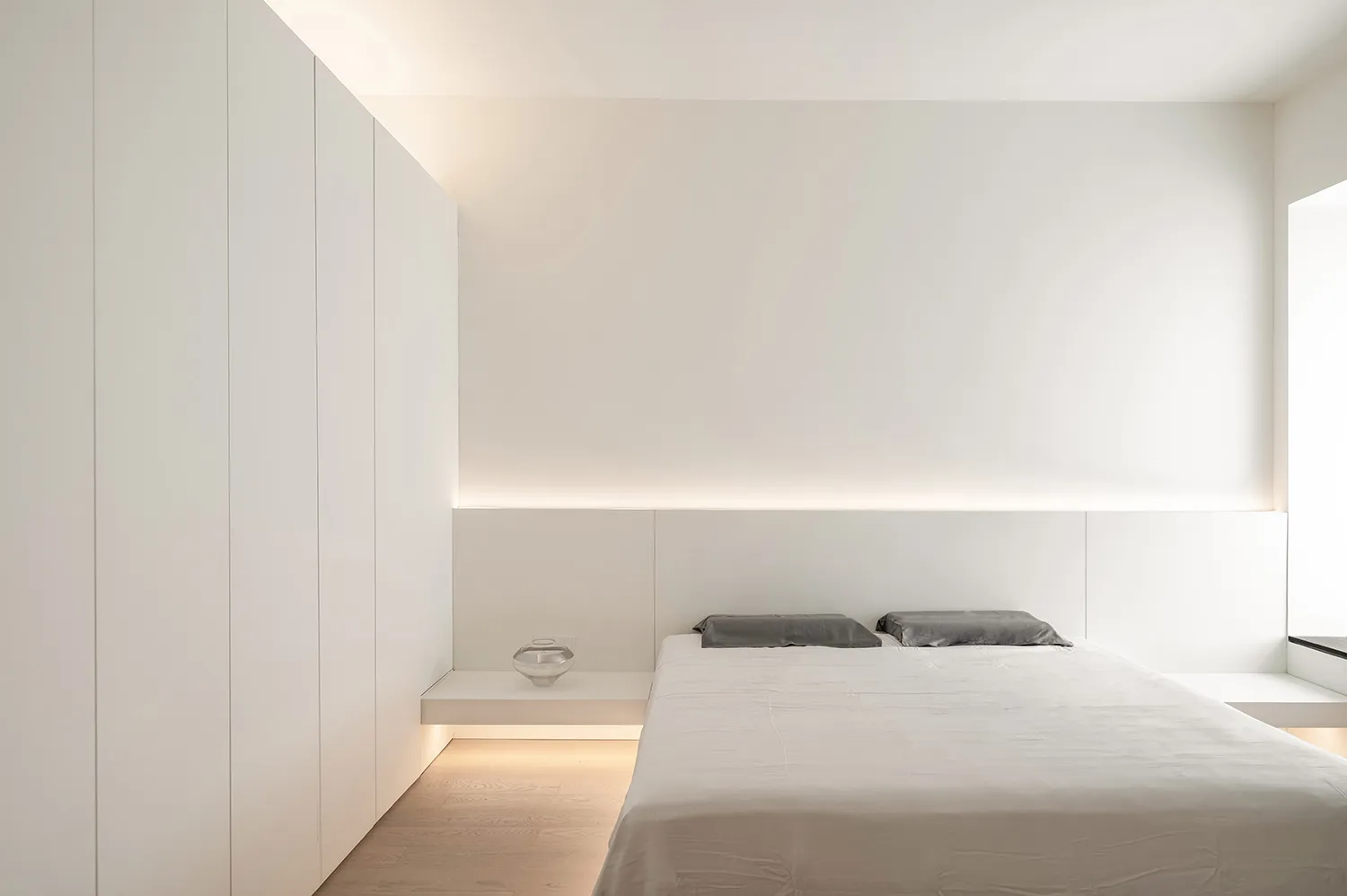 Modern minimalist bedroom featuring a white wall with built-in shelves, a floating bed frame, and grey bedding.