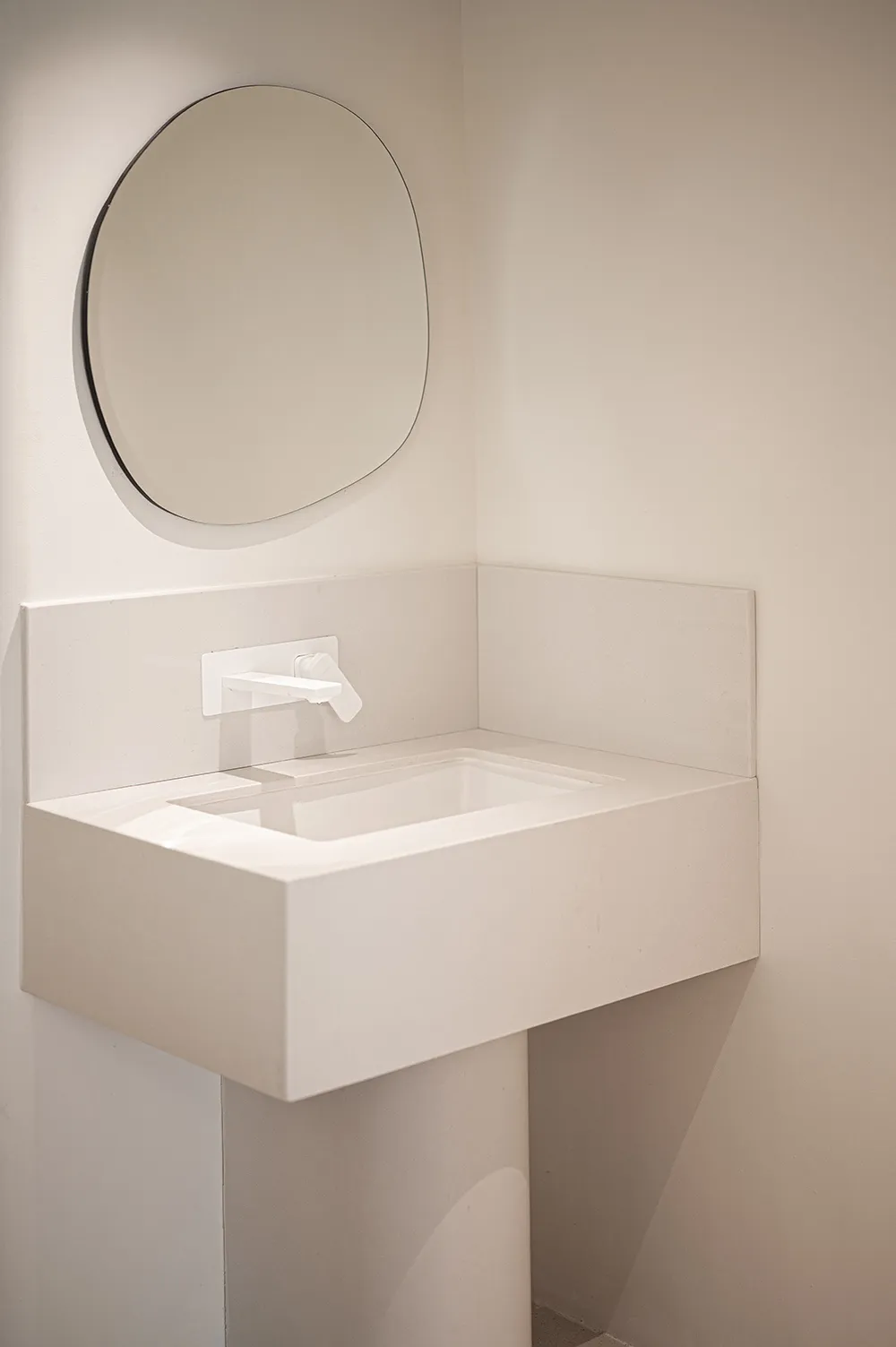 A modern minimalist bathroom with a white sink, concrete counter, and a round mirror on a white wall.