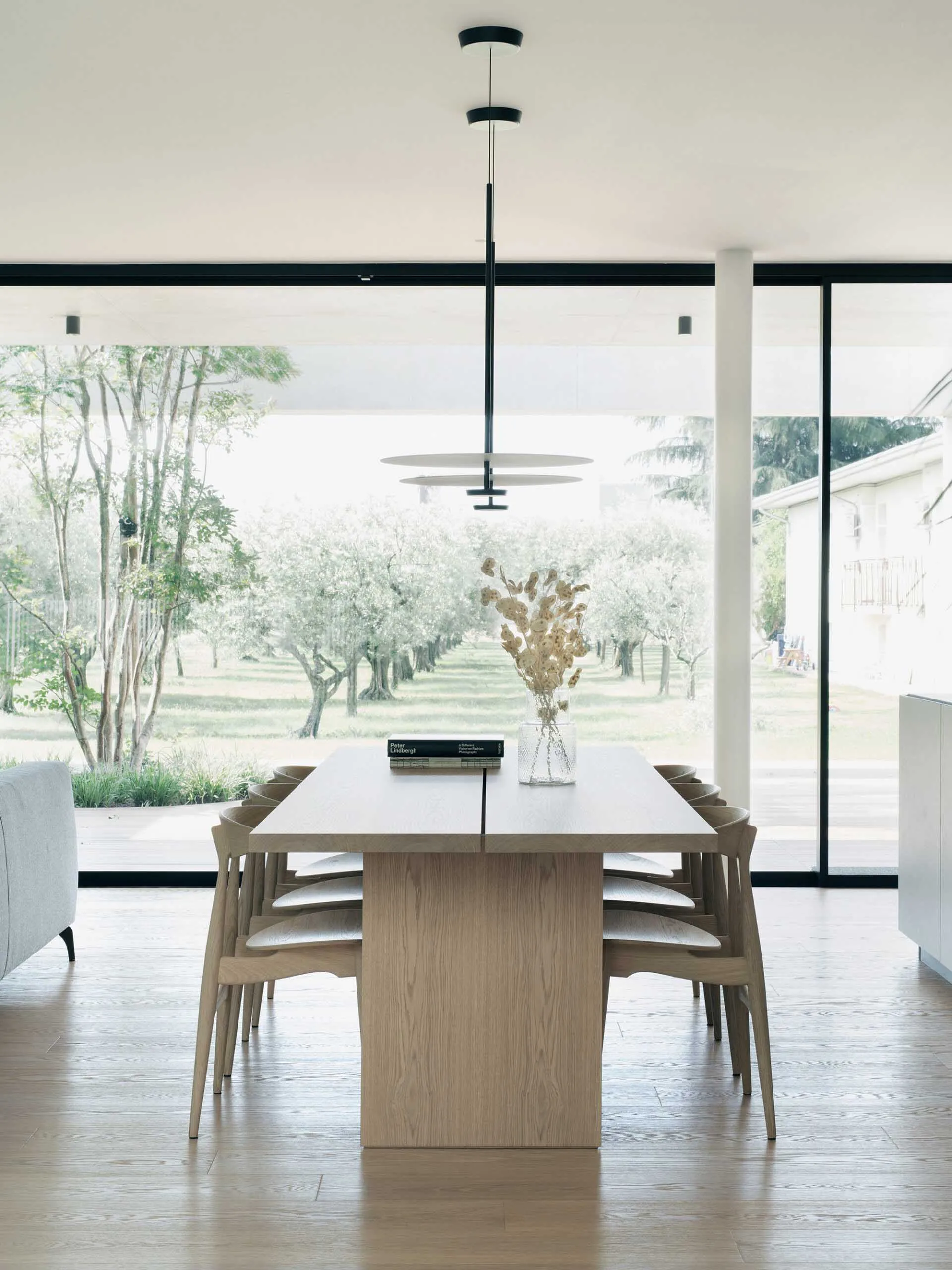 Modern minimalist dining room featuring a wooden table and chairs, with floor-to-ceiling windows showcasing an outdoor olive