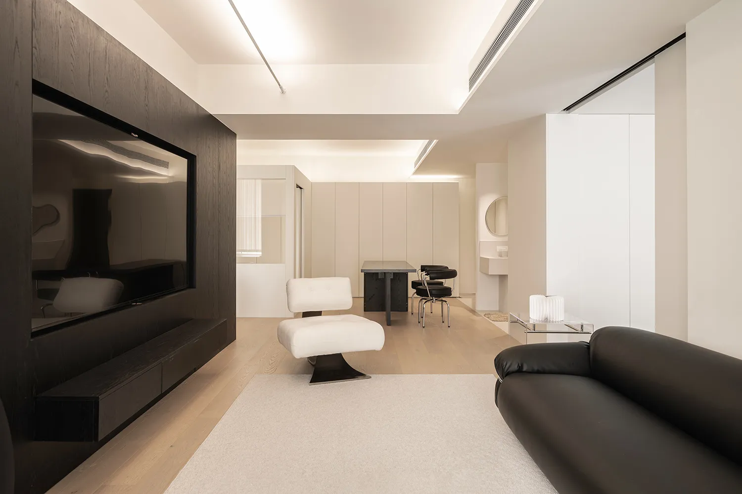 Modern, minimalist living room featuring white and neutral-toned decor with a wooden floor, white rug, black sofa, and white