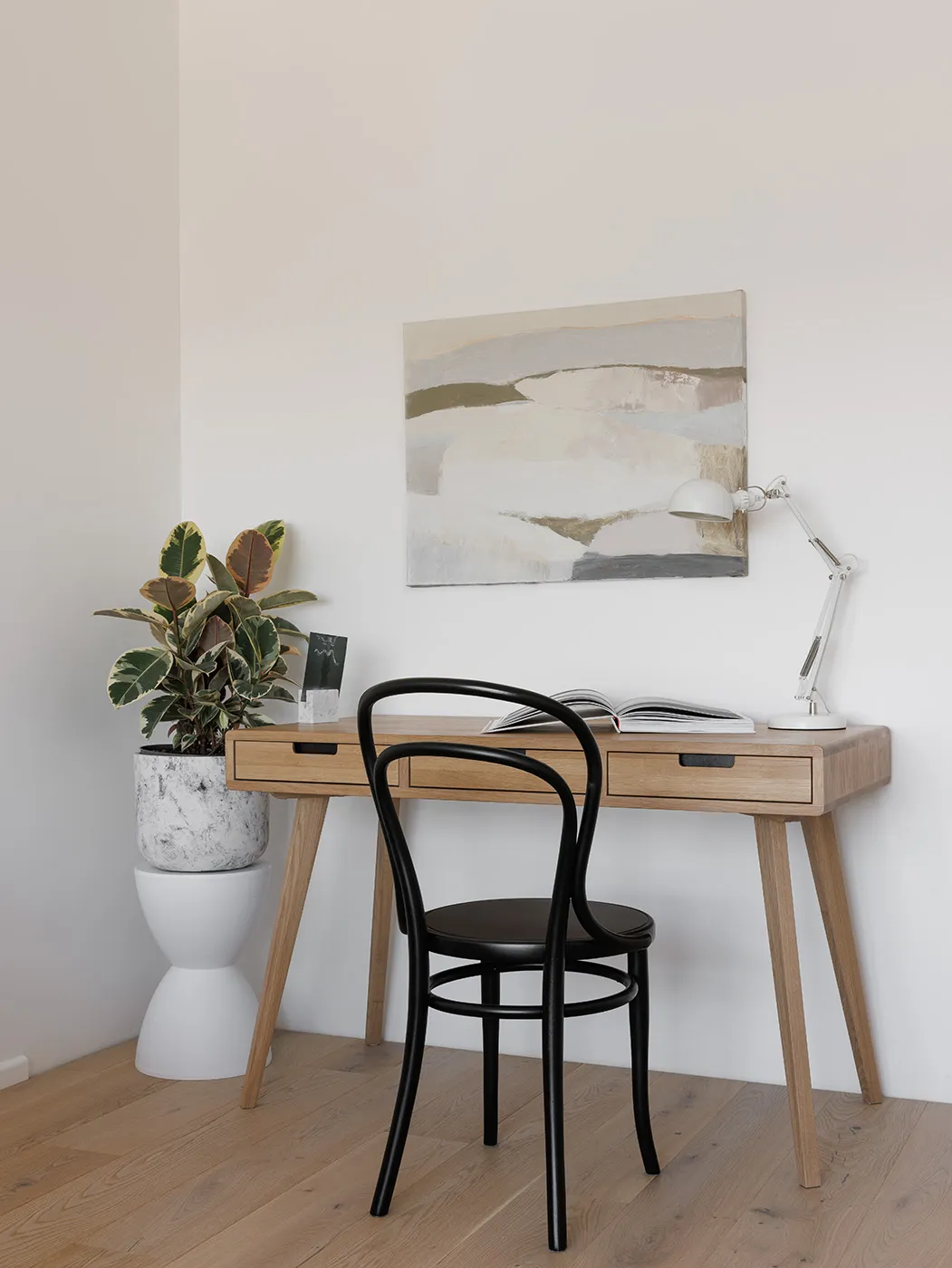 A modern work station featuring a wooden desk with a marble planter and a black chair, with a lamp and an abstract painting.