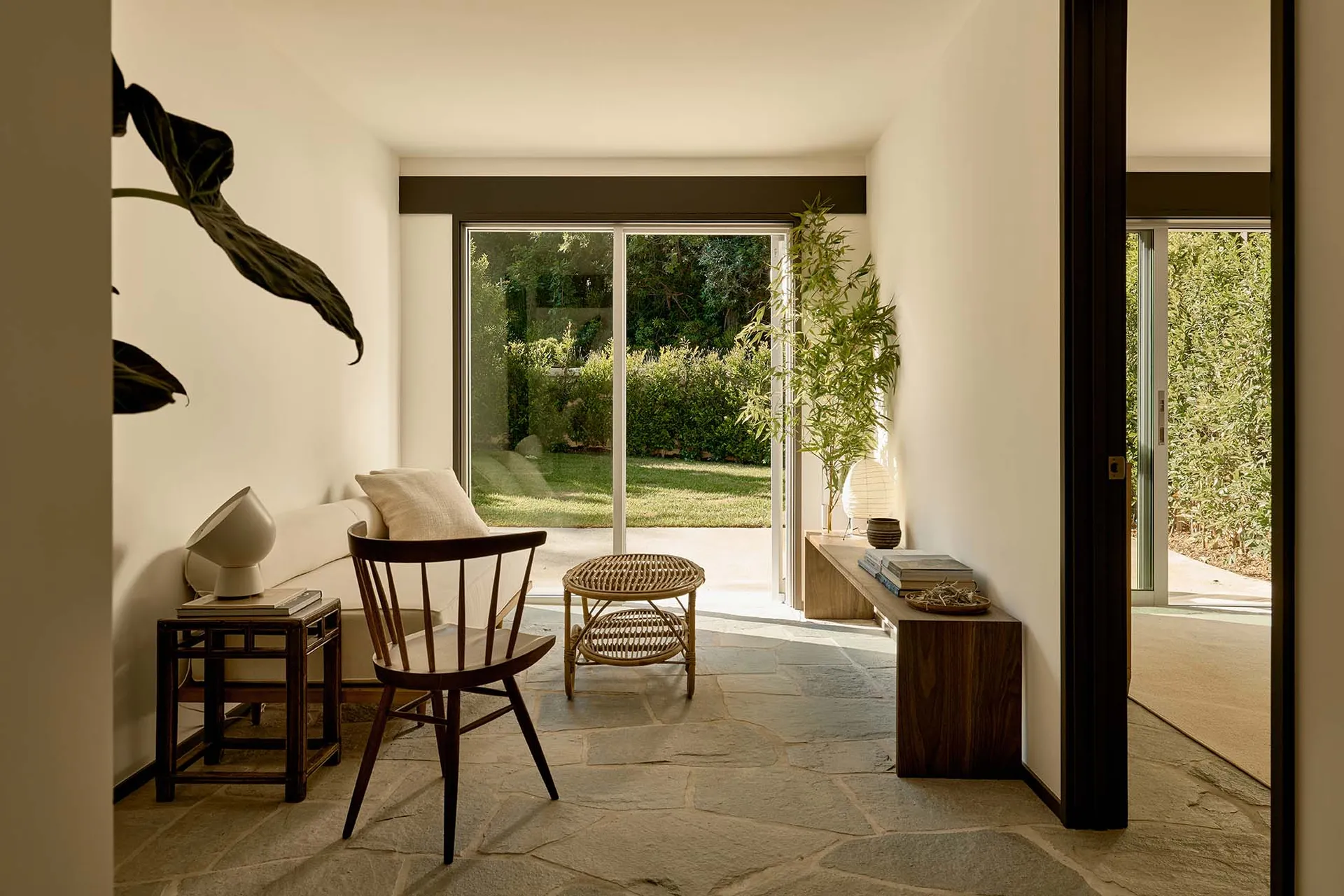 A minimalist living room featuring a stone floor, a white couch, a wooden chair, and a bamboo plant.
