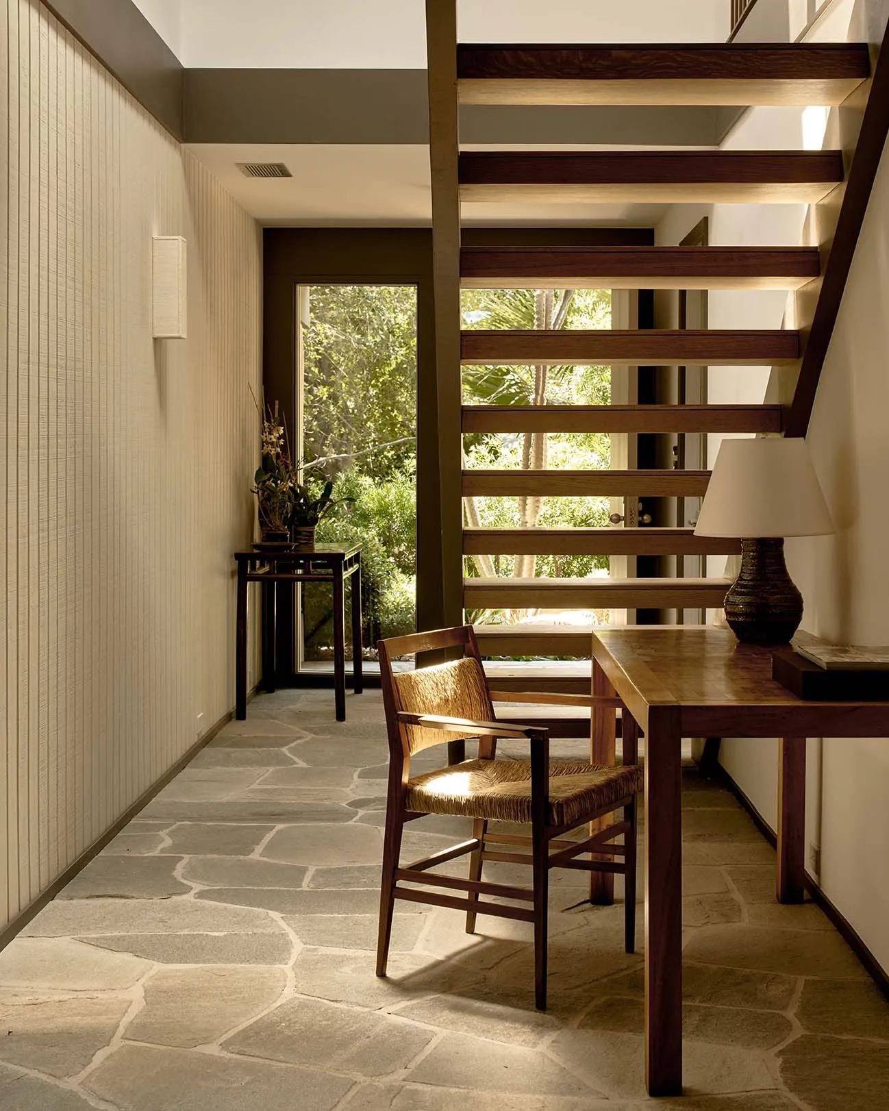 A modern home office with wooden furniture and stone flooring, featuring a built-in desk, chair, and large window.