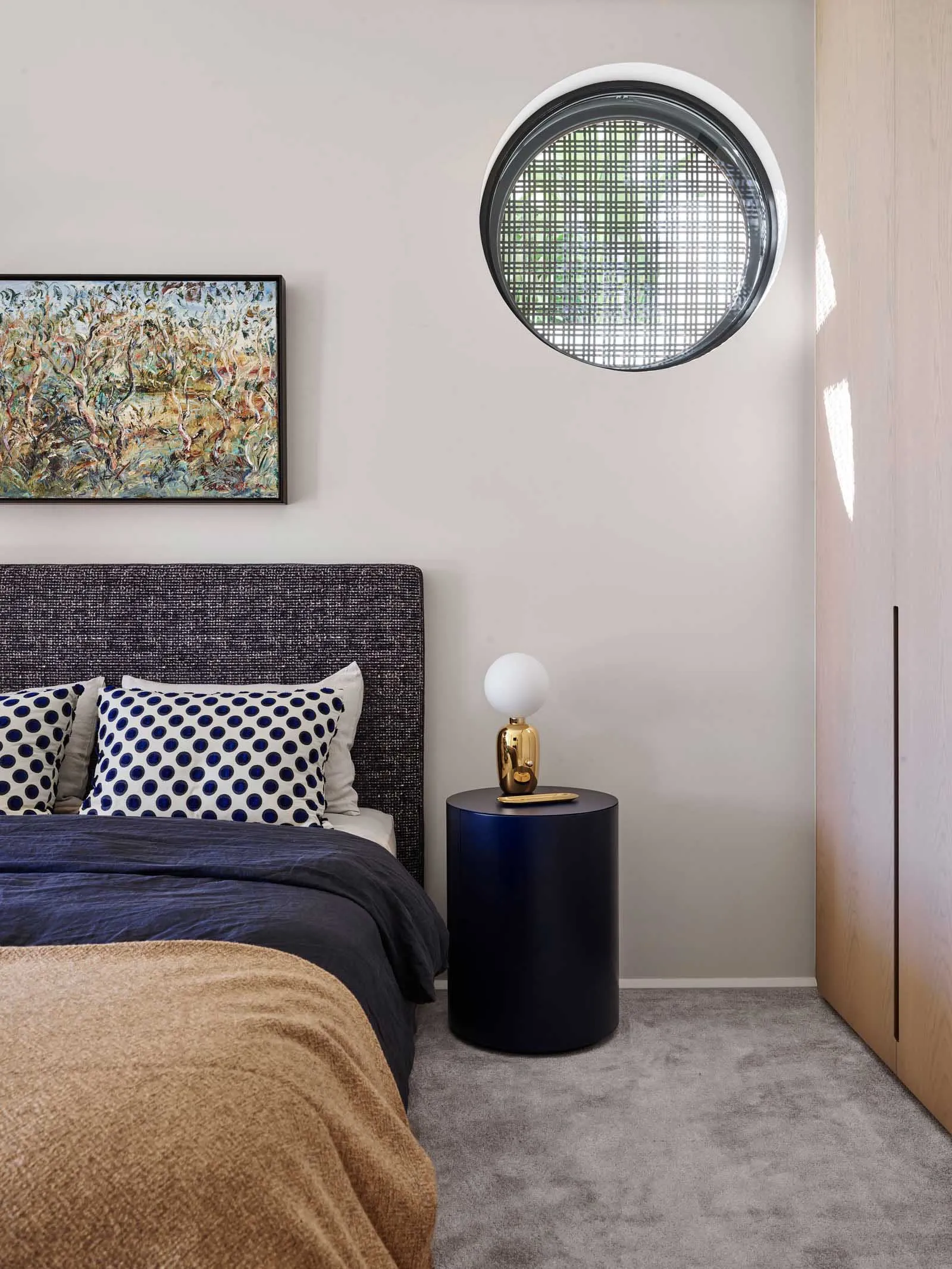 A minimalist bedroom with a round window, fabric headboard, and glass bedside table.