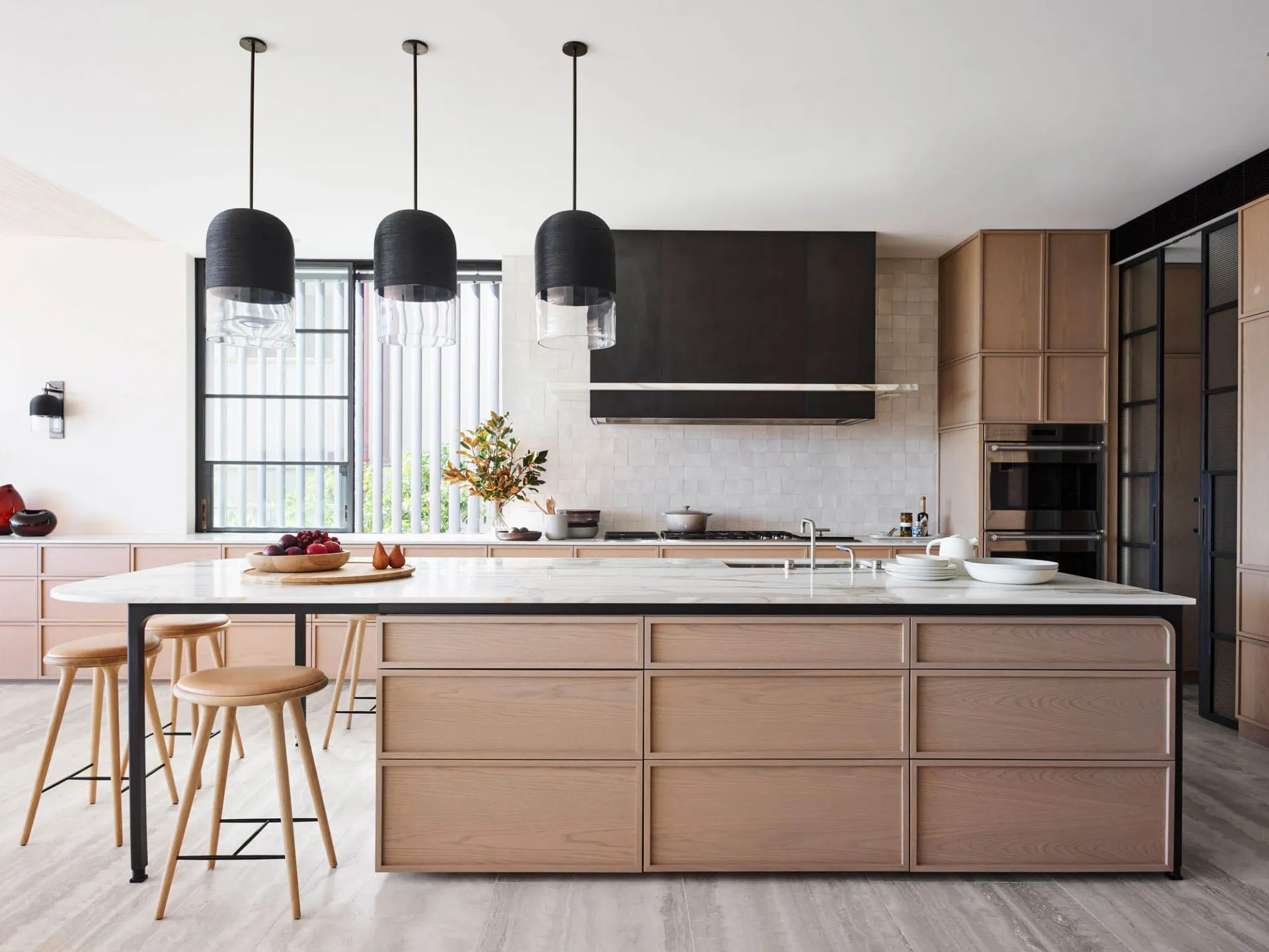 A modern residential kitchen featuring a large island with marble countertop and wooden drawers. The kitchen includes a backs