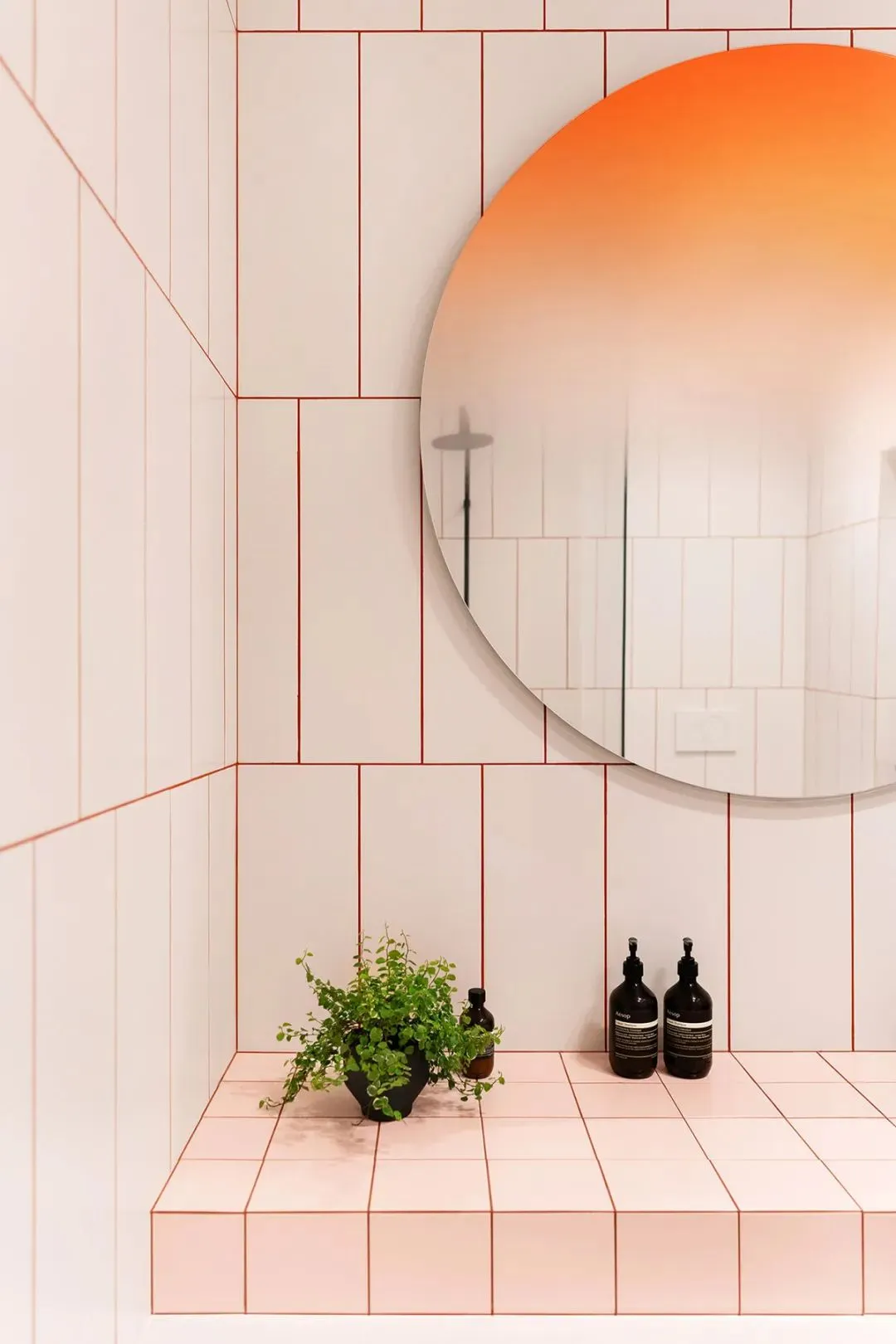 Circular mirror with orange gradient reflection, white ceramic tiles with red grout lines, plant, and two black bottles on ti