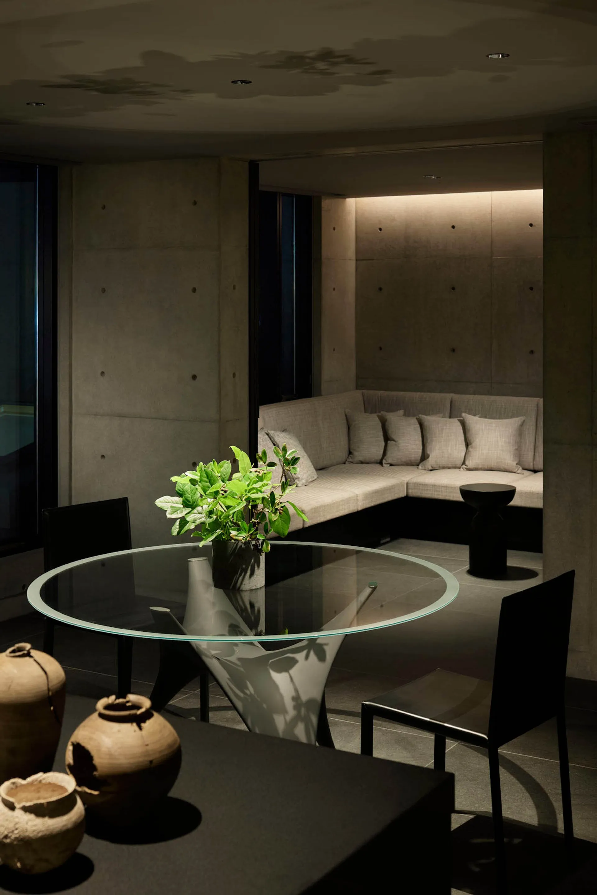 A minimalist living room featuring a concrete wall, circular glass table, and black chairs.