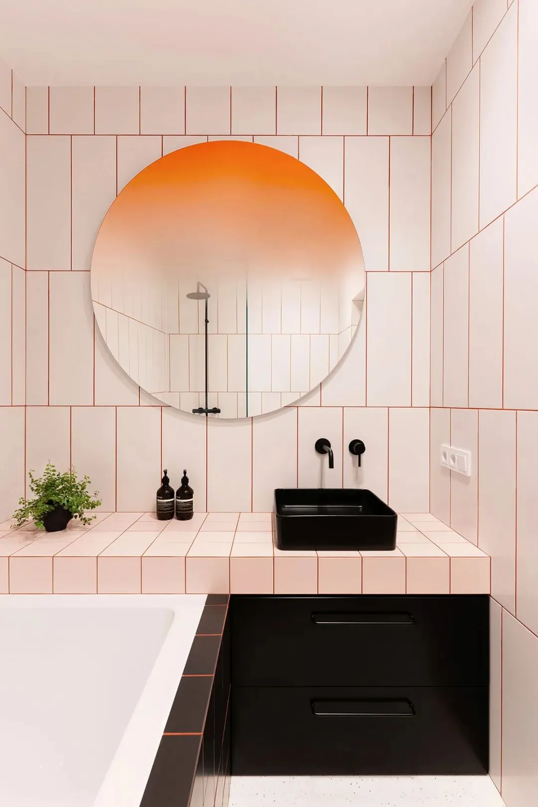 A modern bathroom with white ceramic tiles, a black sink, and an orange gradient circular mirror.